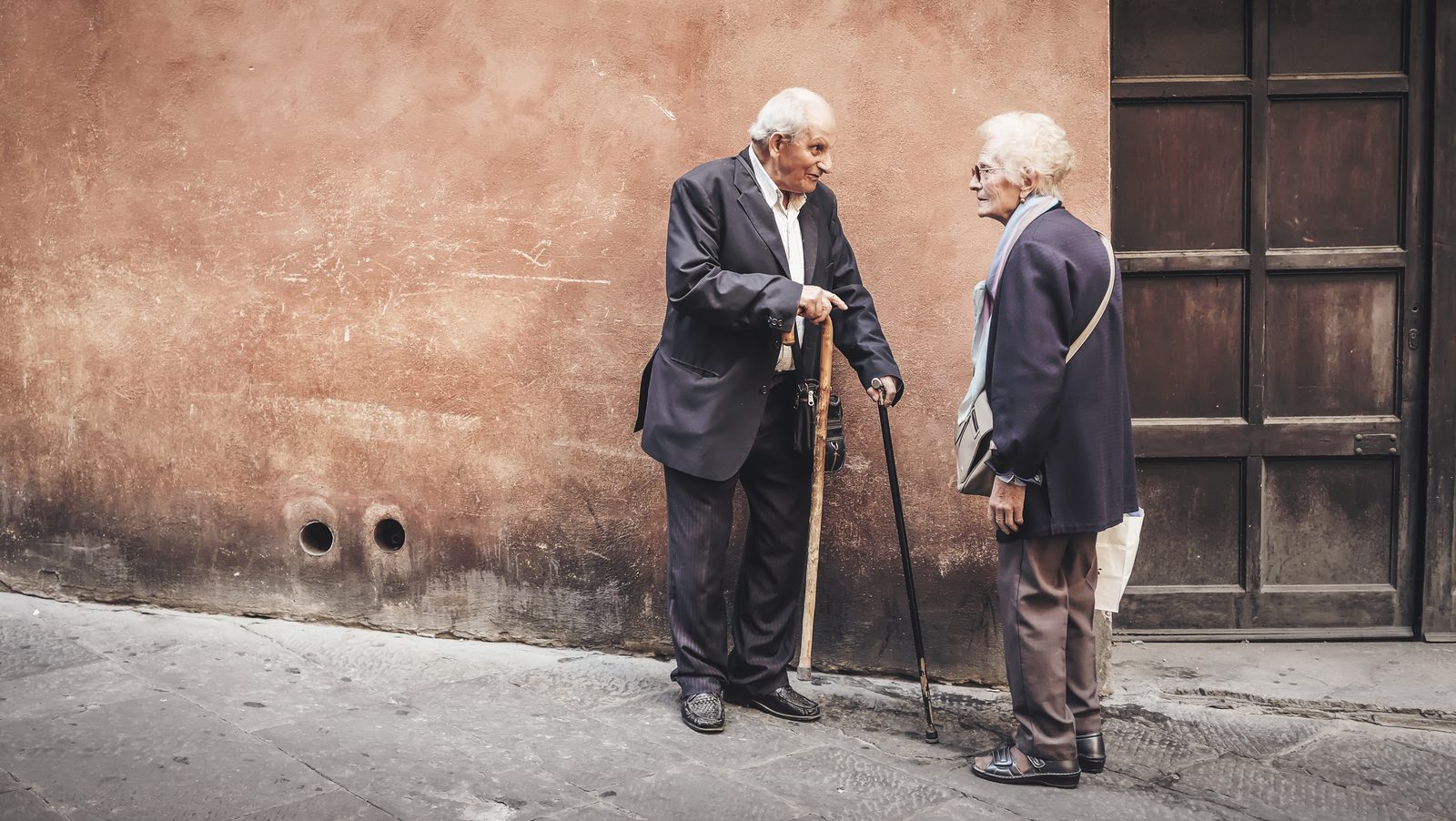 An older man and woman standing outdoors near a doorway, conversing while holding walking sticks.