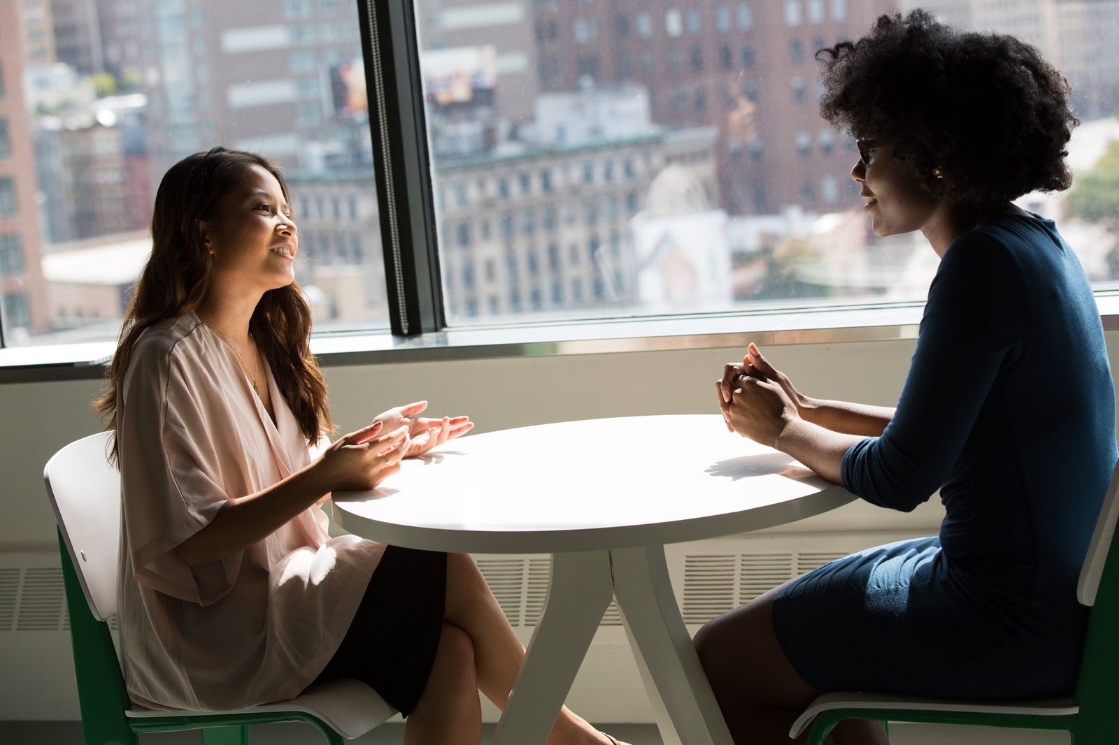 Two women sitting at a table outdoors, talking face to face against a city backdrop.
